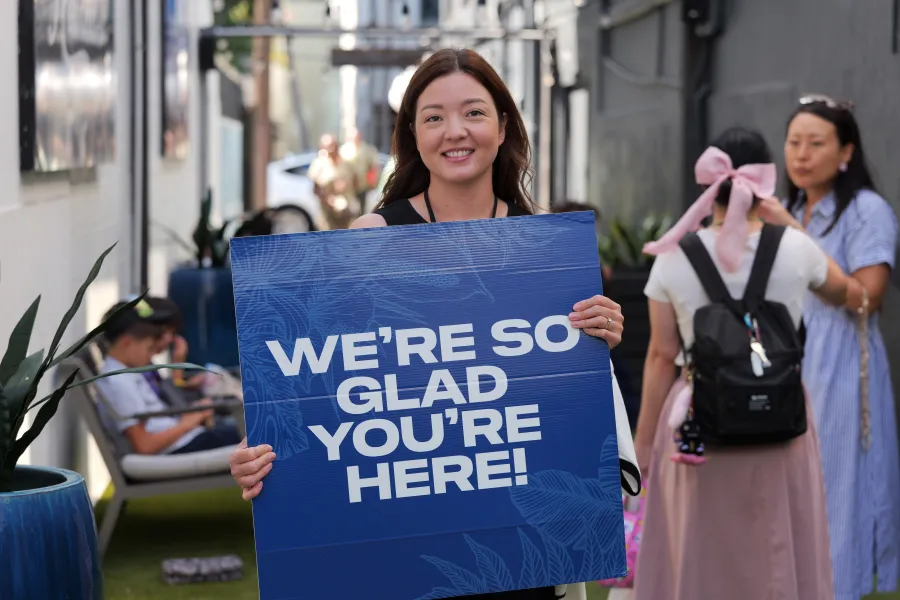A Huikala greeter smiling and holding a sign that reads We're so glad you're here!